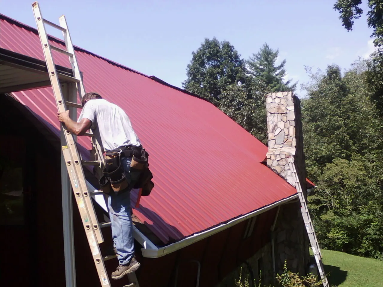 Roofer on a ladder beside a red metal roof and stone chimney — Soffit & Fascia Repair by Roofing Brothers in Crimora